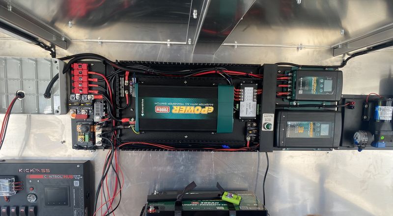 The twelve volt wall of a trailer: a 2000 watt inverter, breakers, fuse blocks and two charge monitors wired above a bank of lithium batteries.