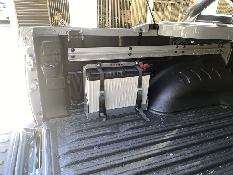 An auxiliary battery strapped into its bracket against the bulkhead of a ute tub, wired and kept clear of the load space.